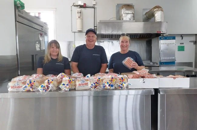 Three people smiling at the camera with blue shirts on and bread laid out in front of them