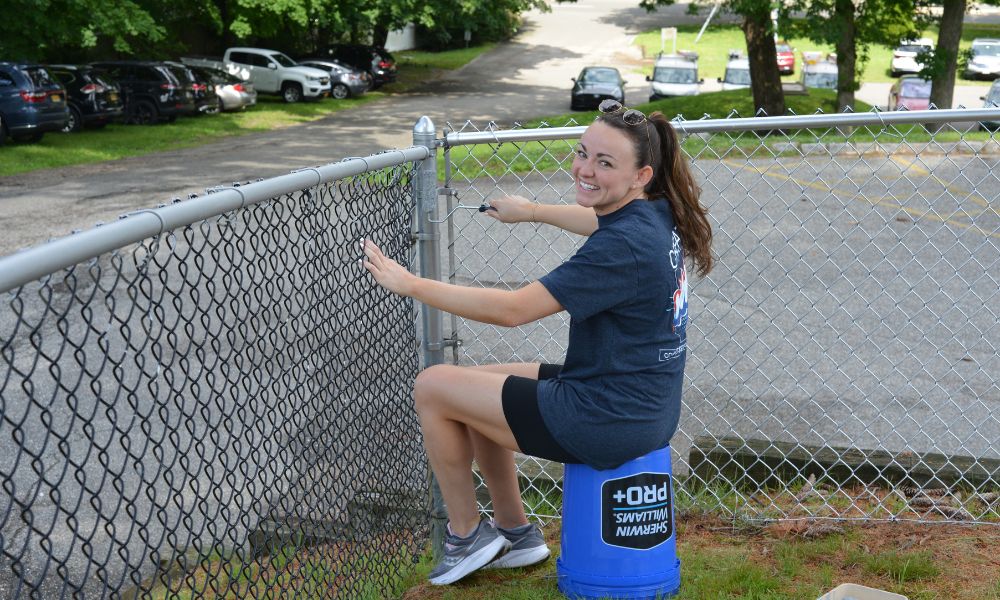 A woman with dark hair in a ponytail and a blue shirt sitting on a blue bucket painting a chain link fence